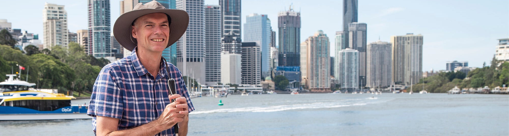 Man standing by a waterfront with a city skyline in the background. The man is holding a white cane.